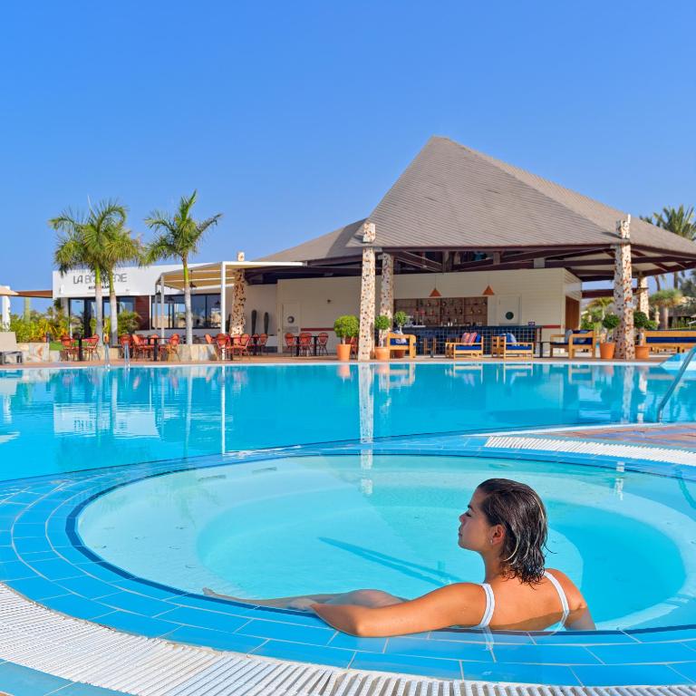 Skaista baseina aina ar relaksējošu siltā ūdens burbuļvannu, kurā atpūšas jauna sieviete. Fonā ir tropu ainava un mūsdienīga atpūtas zona.