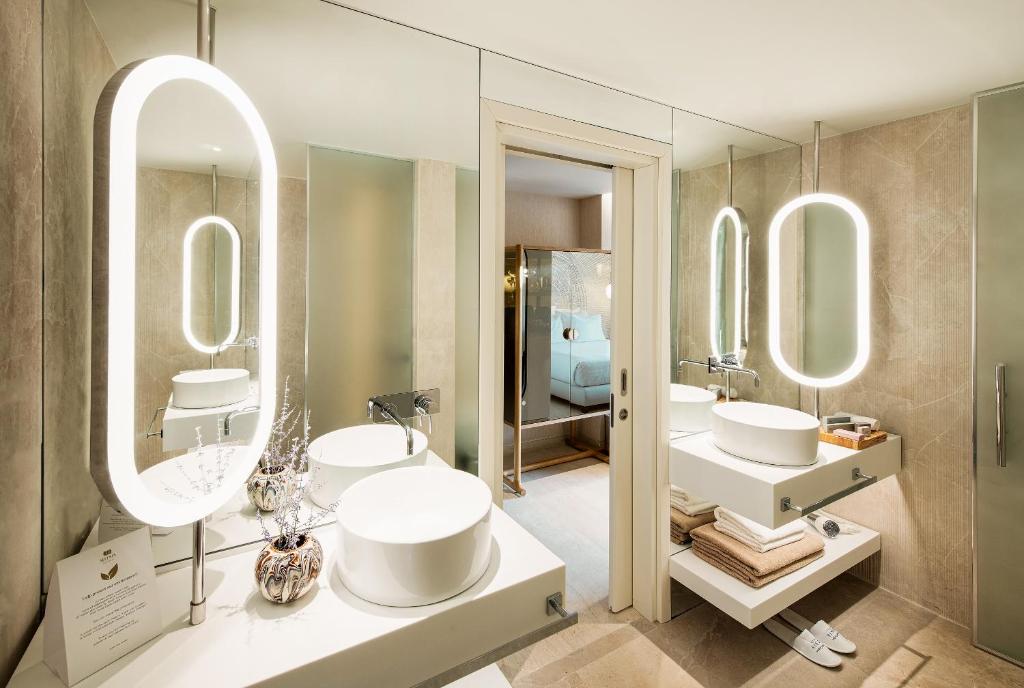 Mūsdienīgs vannas istabas interjers ar apgaismotiem spoguļiem, elegantu izlietni un minimālistisku dizainu. Svaigi salikti dvieļi un dekoratīvi elementi piešķir telpai stilīgu izskatu.