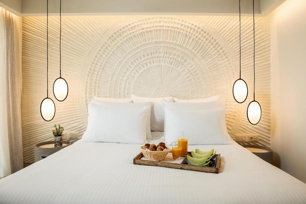 Moderns guļamistabas interjers ar elegantu gultu un stilīgu apgaismojumu. Uz gultas atrodas brokastu šķīvis ar augļiem un dzērieniem.