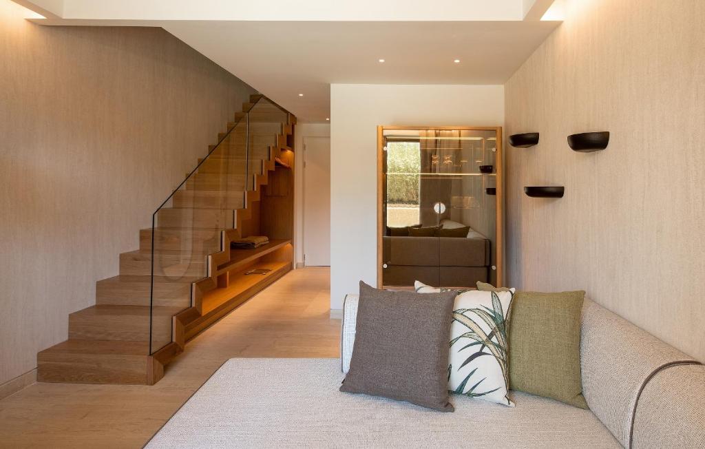 Mūsdienīga interjera telpa ar elegantu koka kāpnēm, stikla margām un mīksto dīvānu, rotātu spilveniem. Gaismas un siltas krāsas rada mājīgu atmosfēru.