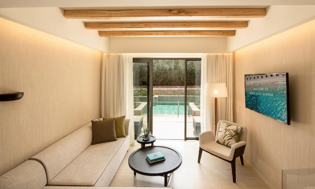 Mūsdienīgs viesistabas interjers ar dabīgiem koka stieņiem, gaišām krāsām un skatu uz baseinu. Ērta dīvāna un krēsla komplekts, kas rada relaksējošu atmosfēru.