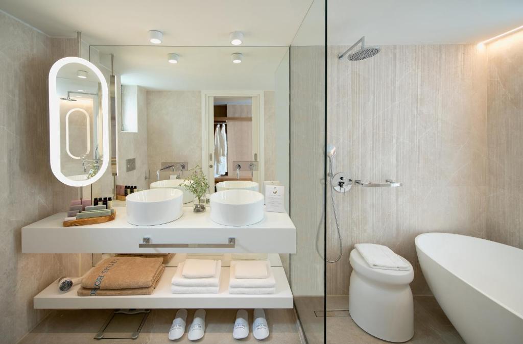 Mūsdienīga vannas istaba ar divām baltām izlietnēm, elegantu apgaismojumu un minimālistisku dizainu. Interjers ir veidots ar neitrālām krāsām un modernām iekārtām, radot relaksējošu atmosfēru.