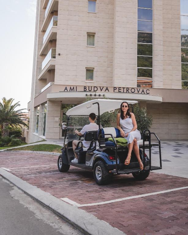 Sieviete baltā kleitā sēž uz golfa ratiņiem, kamēr zēns tos vada, fona redzama viesnīca "Ami Budva Petrovac".