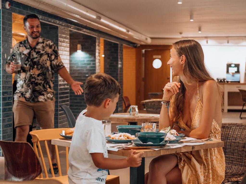 Ģimenes brīvdienu brīdis restorānā, kur pie galda sēž sieviete un bērns, kamēr vīrietis pievienojas ar dzērienu rokā. Interjers ir mūsdienīgs un mājīgs.