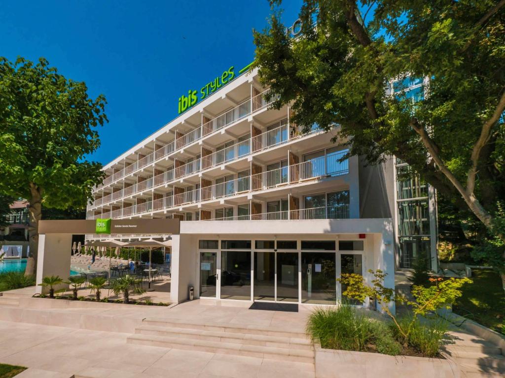 Ibis Styles viesnīcas ēka ar modernu arhitektūru un plašu terasi, apzaļumota ar kokiem un augiem, atrodas saulainā dienā. Galds un krēsli ir izvietoti pie baseina.
