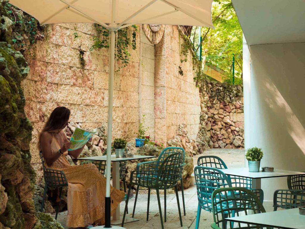 Sieviete sēž kafejnīcā zem jumta, turot rokās karti un izskatoties apkārt. Apkārt ir akmeņu mūri un zaļumi, kas rada mierīgu atmosfēru.