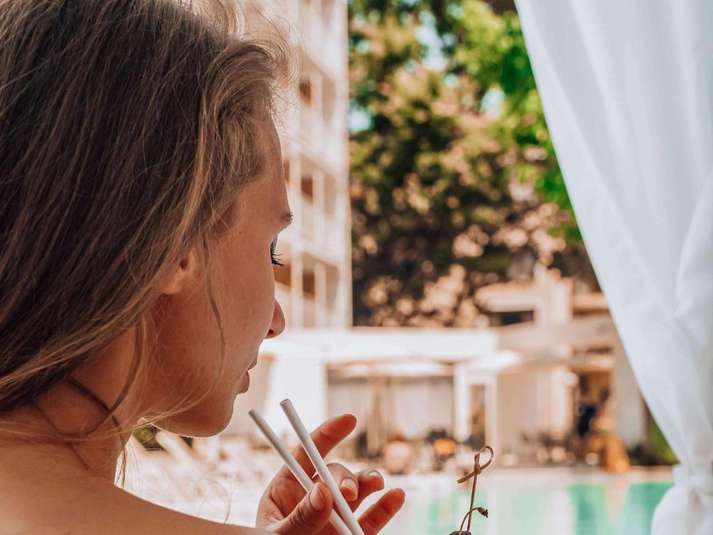 Jauna sieviete relaksējas pie baseina, turēdama rokās divas balto salmiņu. Fons ir saulains un zaļš, radot atpūtas atmosfēru.