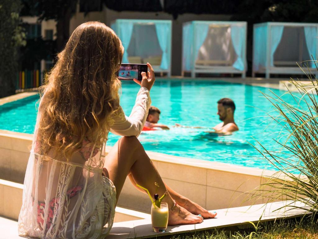 Sieviete ar gariem matiem sēž pie baseina un fotografē divus cilvēkus, kas peld. Blakus viņai ir kokteilis, un fons ir saulains un relaksējošs.