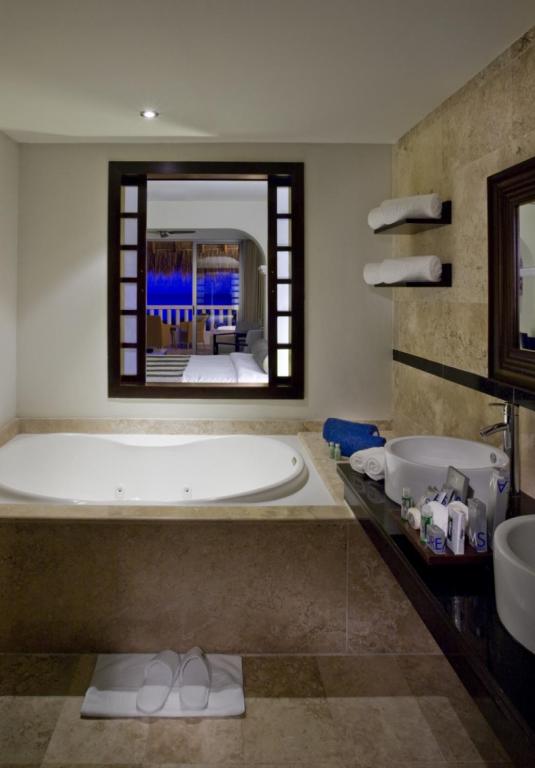 Mūsdienīgs vannas istabas interjers ar lielu vanniņu, mīkstām dvieļiem un elegantiem aksesuāriem. Logā redzama relaksējoša telpa, kas papildina miera atmosfēru.