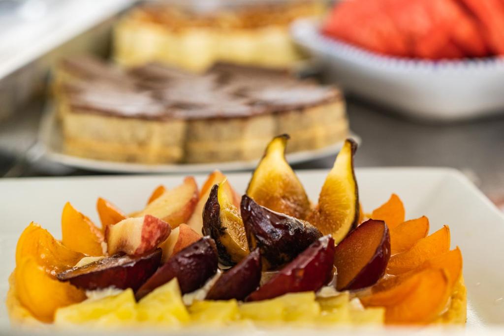 Saldais deserts ar dažādām augļu šķēlītēm, tostarp persikiem un plūmēm, uz balta šķīvja. Fons ietver citus kūkas veidus.