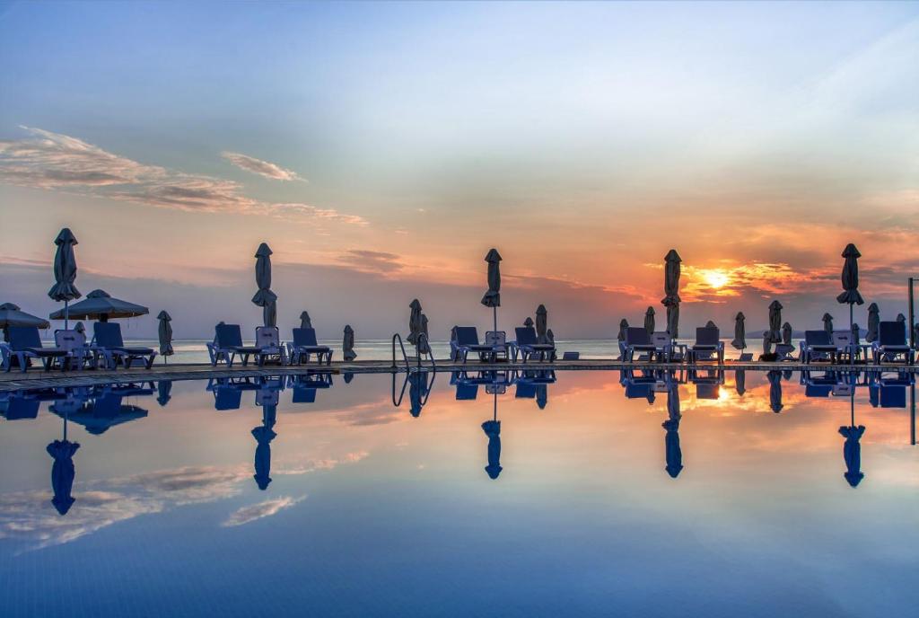 Saule riet, atspoguļojoties mierīgā baseinā, kur atrodas sauļošanās krēsli un saulessargu. Šī ainava rada relaksējošu un idillisku atmosfēru.