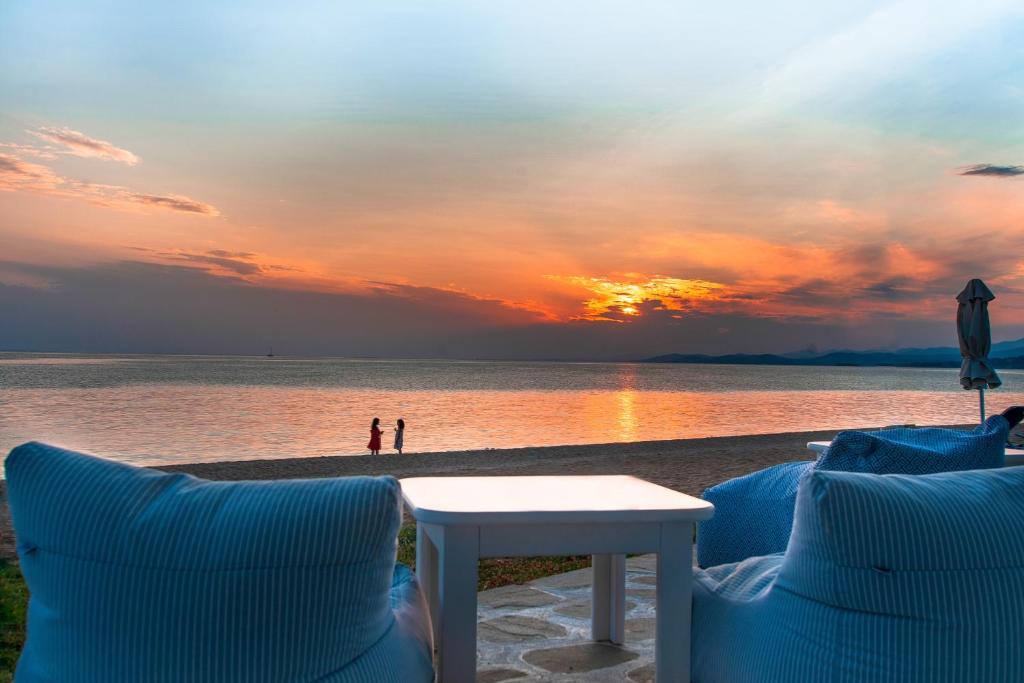 Saule noriet jūrā, radot krāsu spēles debesīs, kamēr divi cilvēki stāv pludmalē, baudot mierīgu vakaru. Priekšplānā redzami ērti krēsli un galds, kas aicina uz relaksāciju.