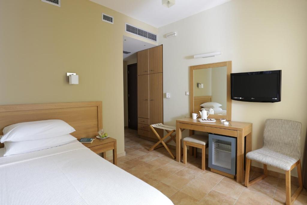 Mūsdienīga viesnīcas istaba ar gultu, rakstāmgaldu un televizoru, iekārtota ar dabīgā koka mēbelēm un neitrālām krāsām. Telpā ir arī ledusskapis un spogulis.