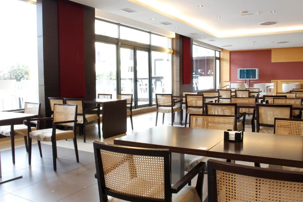 Mūsdienīgs kafejnīcas interjers ar koka galdiem un krēsliem, plašām logu vitrīnām un gaišu apgaismojumu. Telpa ir ērti iekārtota, piedāvājot patīkamu atmosfēru.