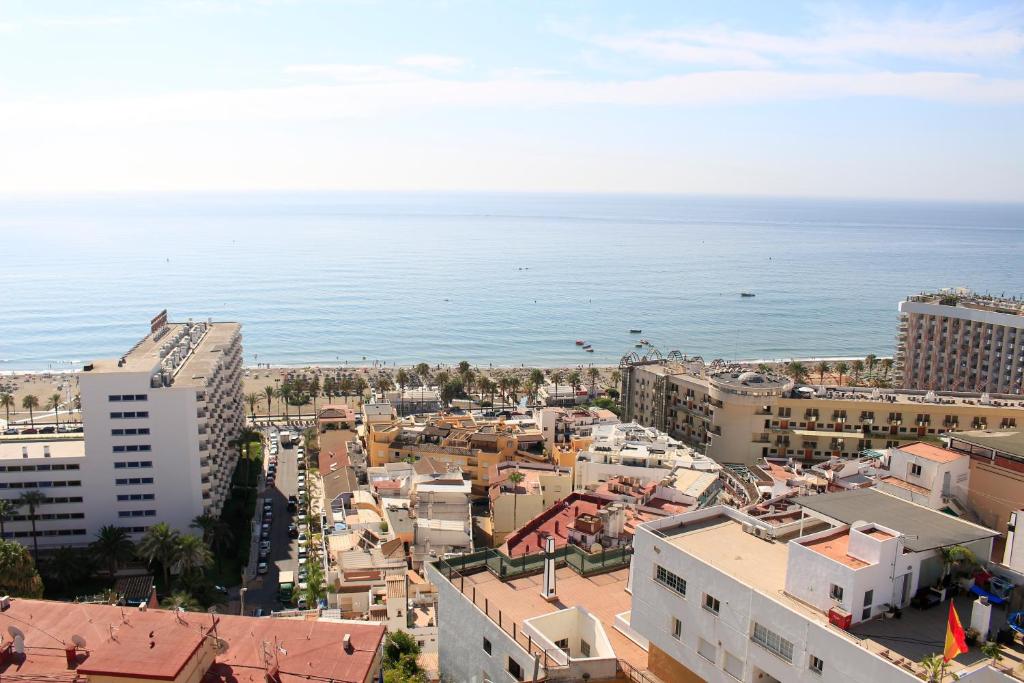 Skats uz pilsētu pie jūras, kur redzami moderni ēku kompleksi un pludmale ar palmām. Aiz ēkām stiepjas mierīgs jūras ūdens un zila debess.