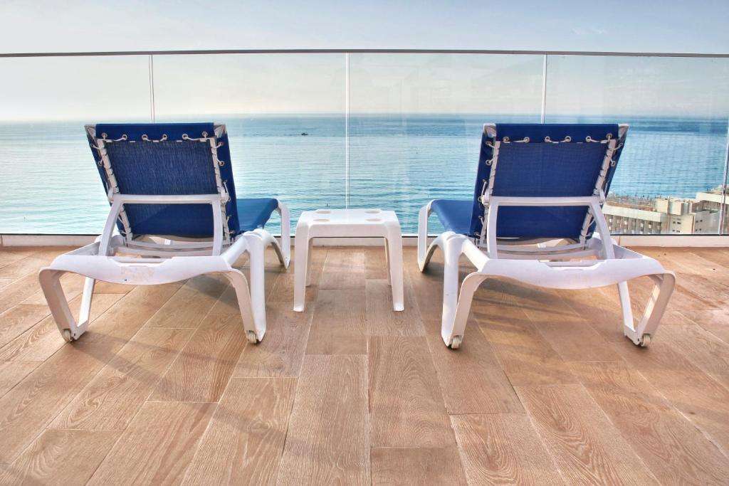 Divi zilu sauļošanās krēsli uz balkona ar skatu uz jūru, starp tiem balts galdiņš. Mierīga atmosfēra un gaiši koka grīdas segums.