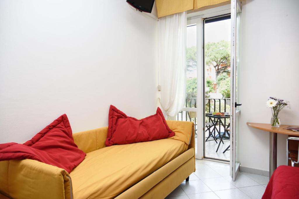 Gaiša viesistaba ar dzeltenu dīvānu un sarkaniem spilveniem, logu uz balkonu, kur redzami dārzi un āra mēbeles. Interjers ir moderns un mājīgs.