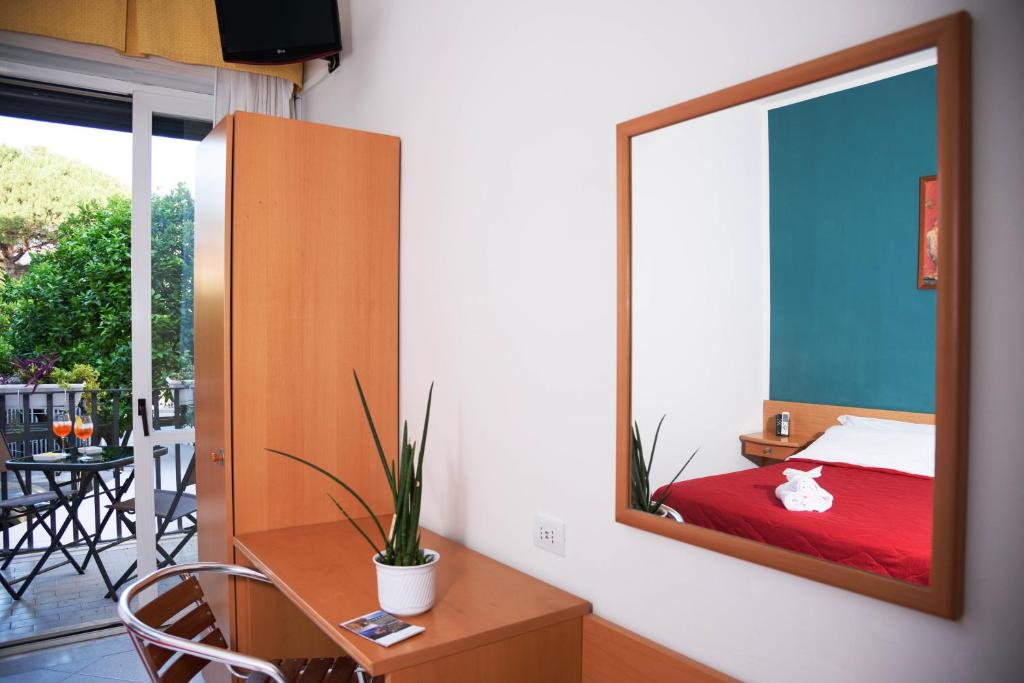 Mūsdienīga viesnīcas istaba ar sarkanu gultas veļu un lielu spoguli, kas atspoguļo ērti iekārtotu telpu. Logā redzama zaļa terase ar galdiem un dzērieniem.