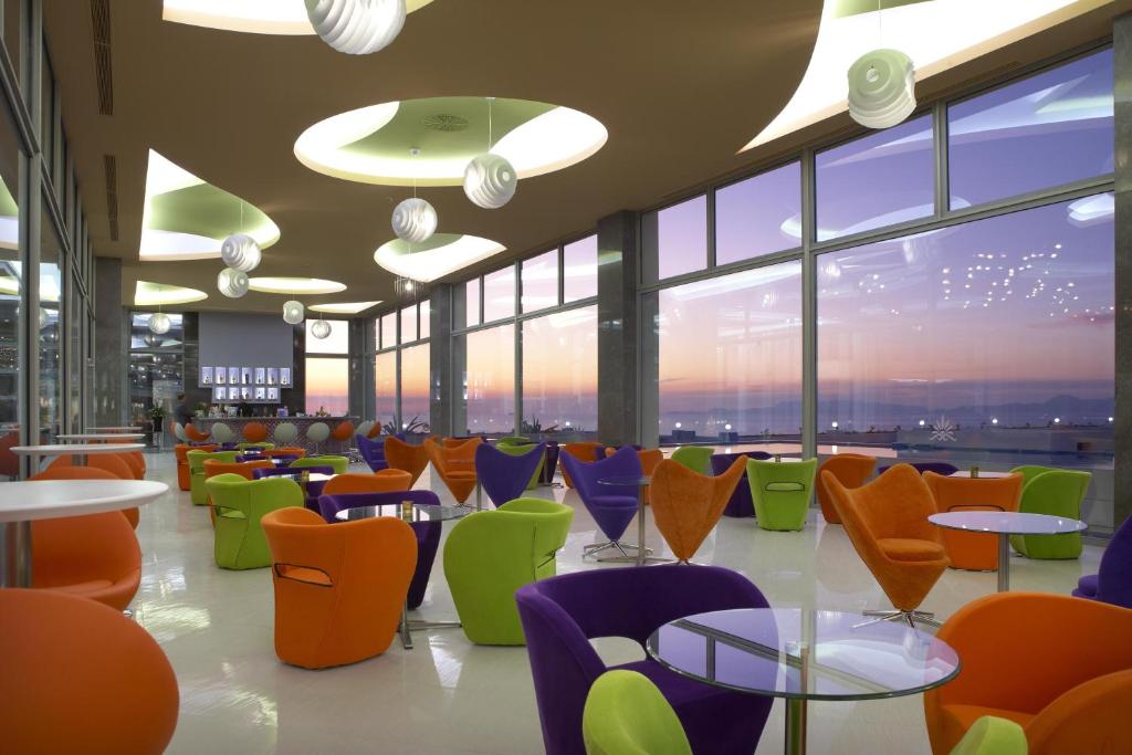 Mūsdienīgs kafejnīcas interjers ar krāsainām mēbelēm un lieliem logiem, caur kuriem redzams saulriets. Sēdvietas ir veidotas no spilgtiem krāsām, radot patīkamu un draudzīgu atmosfēru.