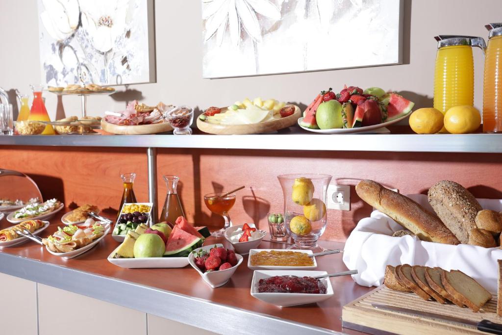 Bagātīga brokastu bufete ar augļiem, maizēm, gaļas izstrādājumiem un dzērieniem, kas izkārtoti uz elegantas plaukta.