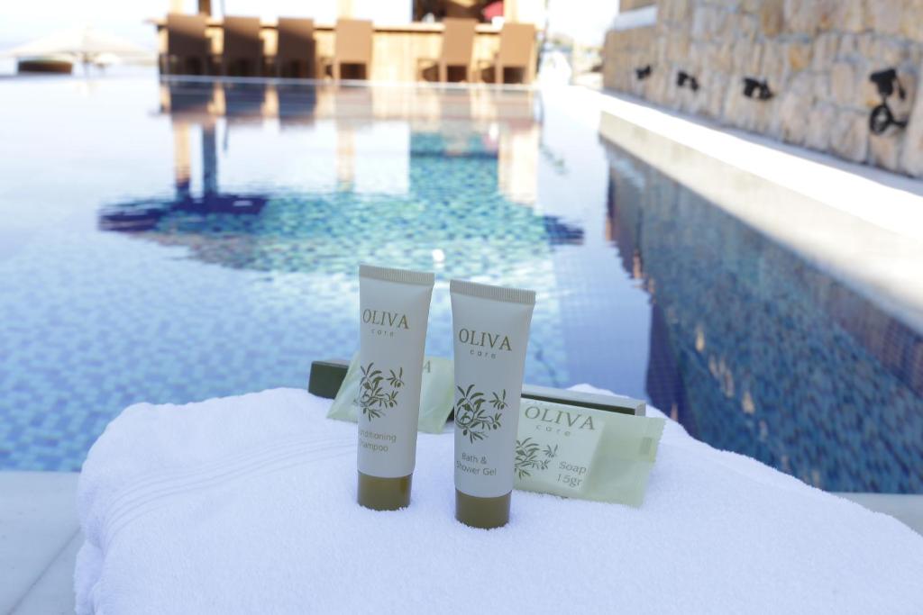 Kosmētikas produktu komplekts ar OLIVA zīmolu, ieskaitot kondicionējošo šampūnu un dušas želeju, novietots uz baltas dvielītes blakus baseinam. Fons veido skaists baseins ar mozaīkas flīzēm un relaksējošu atmosfēru.
