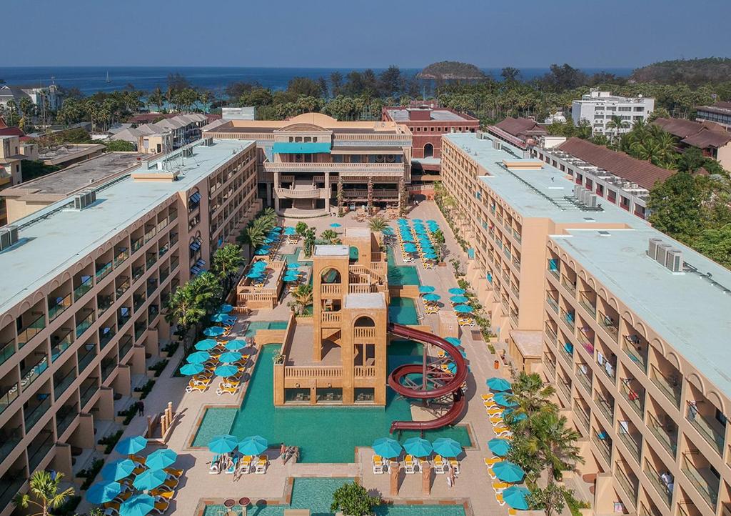 Viesnīcas komplekss ar baseinu un slidkalniņu, kas atrodas piekrastē, apjozts ar palmām un sauļošanās krēsliem. Fons ietver jūru un apkārtējās ēkas.