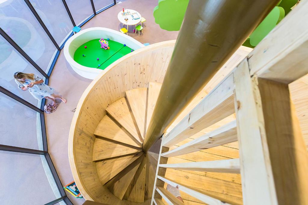 Koka spiralveida kāpnes ved uz bērnu rotaļu zonu ar zaļu paklāju un rotaļlietām. Attēls parāda modernu interjeru ar lieliem logiem, kas nodrošina dabisko apgaismojumu.