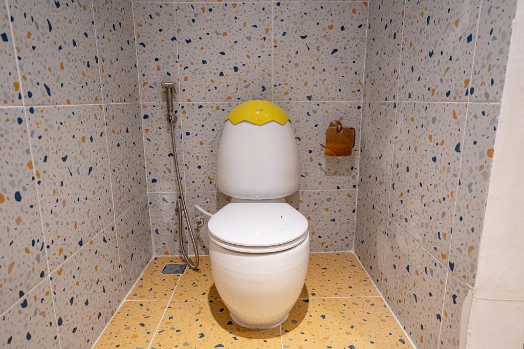 Tualete ar radošu dizainu, kas izskatās kā ola, ar dzeltenu vāciņu un flīzēm ar krāsainiem akcentiem. Interjers ir moderns un stilīgs.