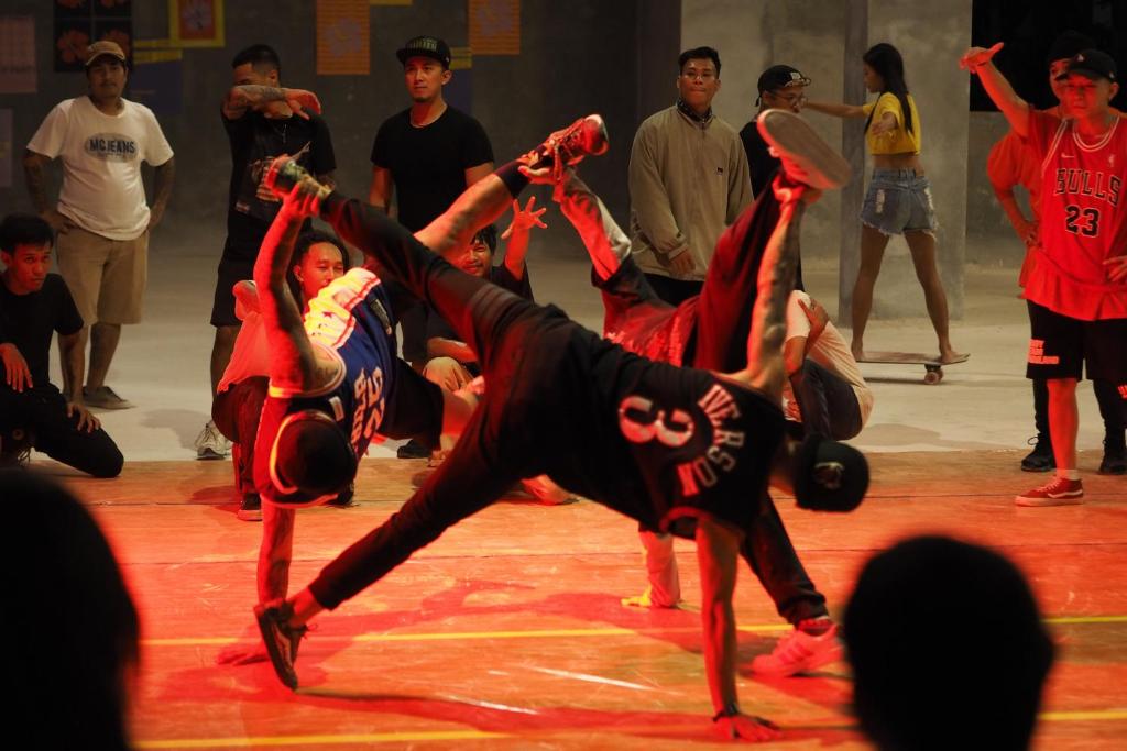 Dejotāju grupas izpilda akrobātiskus dejas soļus, kamēr skatītāji vēro pasākumu. Skatuve ir apgaismota ar sarkanu gaismu, radot dinamisku atmosfēru.