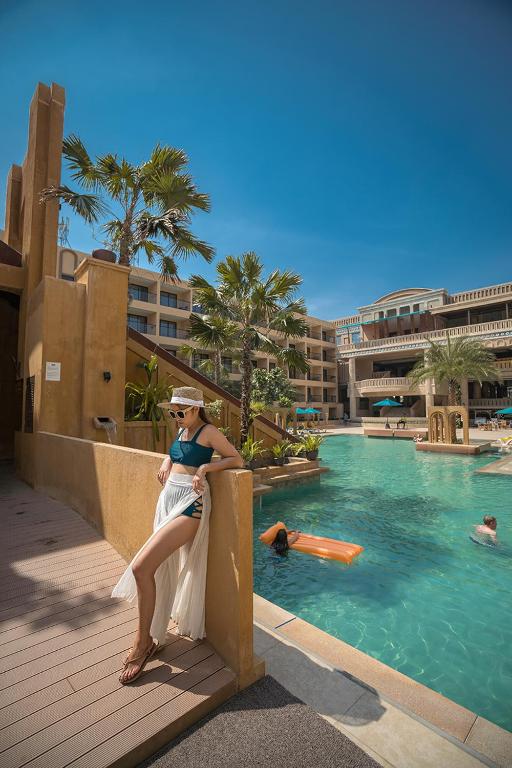 Vasaras brīvdienu atmosfēra pie baseina ar palmām un viesnīcu fonā, kur sieviete apģērbta peldkostīmā un balta svārku daļa.