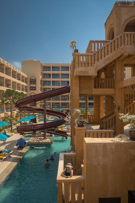 Baseina zona ar ūdens slidkalniņu un viesnīcas ēku fona. Viesi izbauda atpūtu pie ūdens, kamēr saule spīd debesīs.