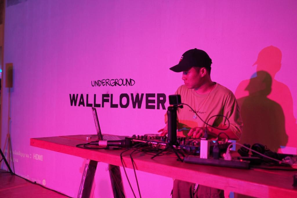 DJ uzstāšanās laikā pie galda ar aprīkojumu, fona siena ar uzrakstu 