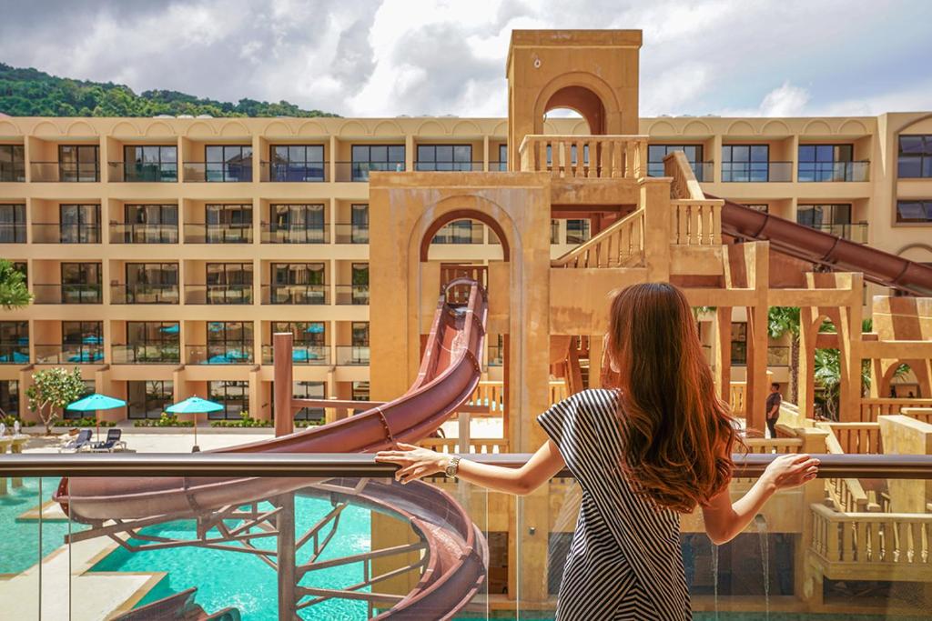 Sieviete ar gariem matiem stāv uz balkona, skatoties uz viesnīcas baseinu ar ūdens slidkalniņu un apkārtējiem viesnīcas numuriem. Aiz viņas ir zils debesis ar dažiem mākoņiem.