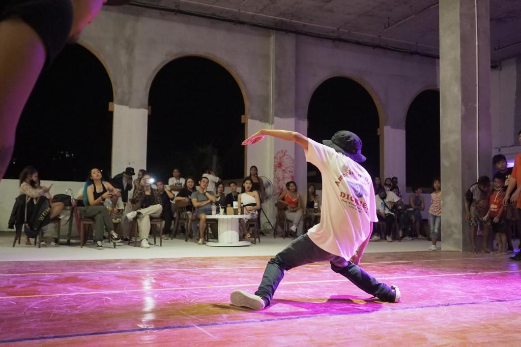 Dejotājs izpilda dinamisku soli uz skatuves, kamēr skatītāji vēro no malas. Pasākums notiek nakts gaismā, radot dzīvīgu atmosfēru.