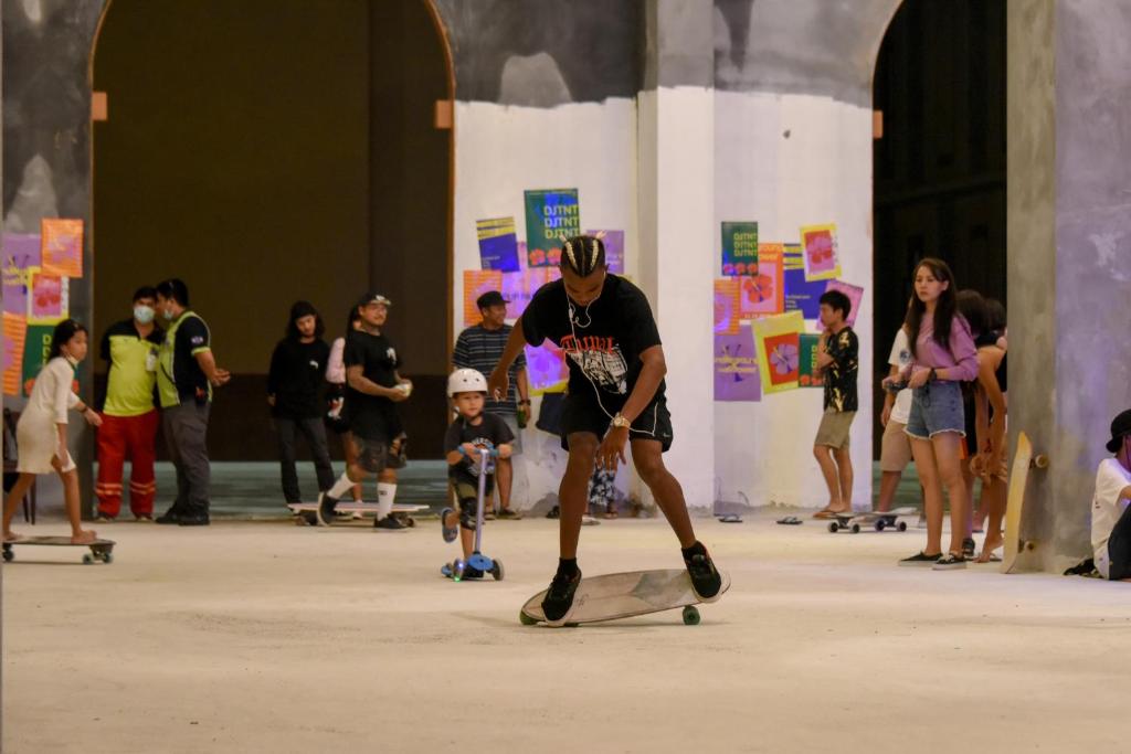 Skatbordotāju pasākums iekštelpās, kur jaunieši un bērni izmēģina savus prasmes uz skatu plāksnēm. Fona plakāti piešķir krāsainu atmosfēru.