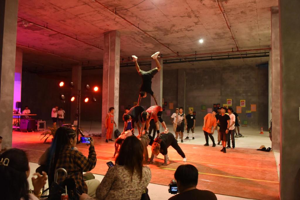 Dejotāju grupa izpilda akrobātiskus trikus, kamēr publika vēro priekšnesumu. Pasākums notiek telpā ar neapstrādātu betona interjeru un sarkanām gaismām.