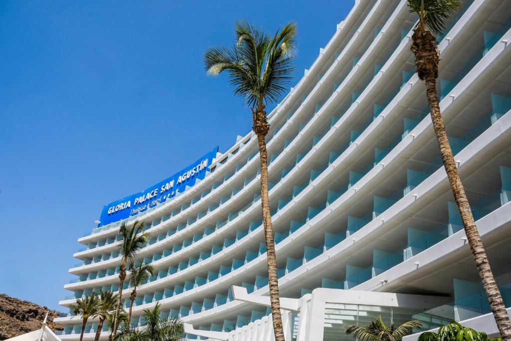 Mūsdienīgs viesnīcas "Gloria Palace San Agustín" ēkas skats ar palmām un skaidru zilu debesi.