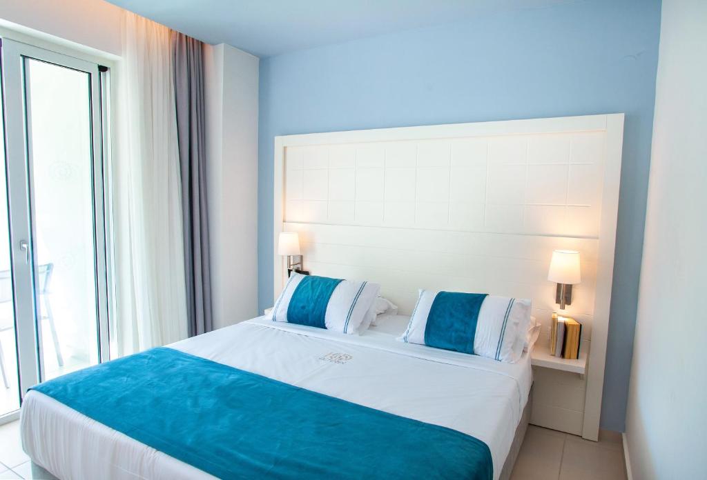 Mūsdienīga guļamistaba ar gultu, baltu sienu un zilu gultas pārklāju, kas radījuši relaksējošu atmosfēru. Lielie logi ļauj ieplūst dabiskajai gaismai un sniedz piekļuvi balkonam.