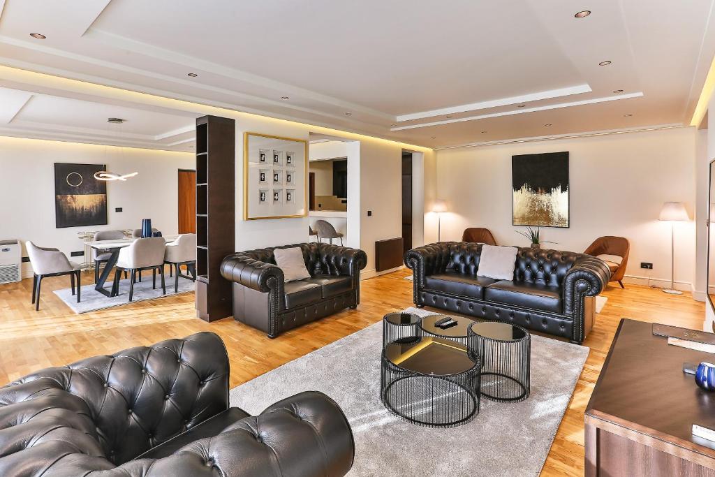 Mūsdienīgs viesistabas interjers ar melnām ādas dīvāniem, stilīgām mēbelēm un koka grīdu. Telpa ir apgaismota ar siltu gaismu un dekorēta ar mūsdienīgām mākslas darba.