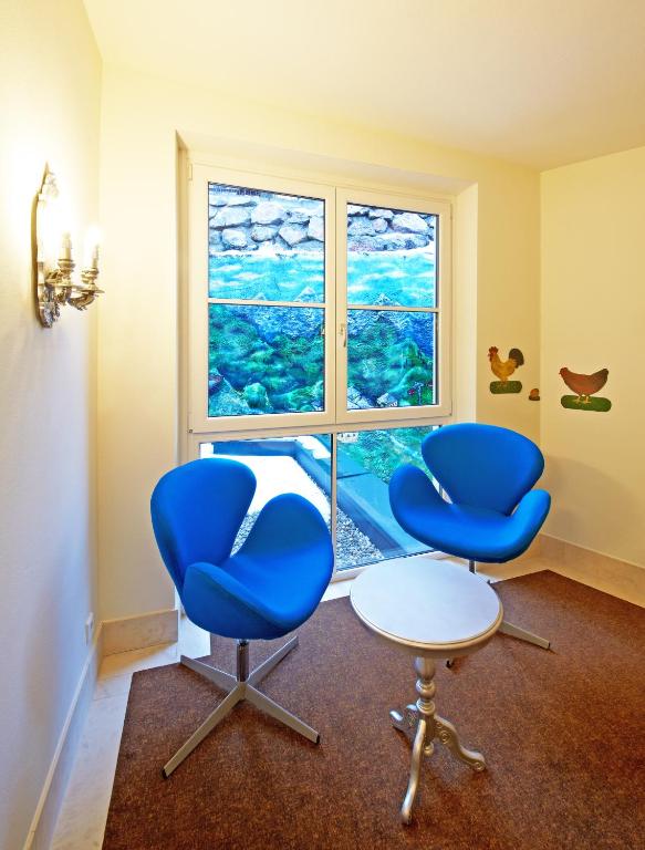 Divas modernas zilas krēsli un mazs galdiņš stūrī, blakus logam ar ainavisku skatu un dekoratīviem elementiem uz sienas.