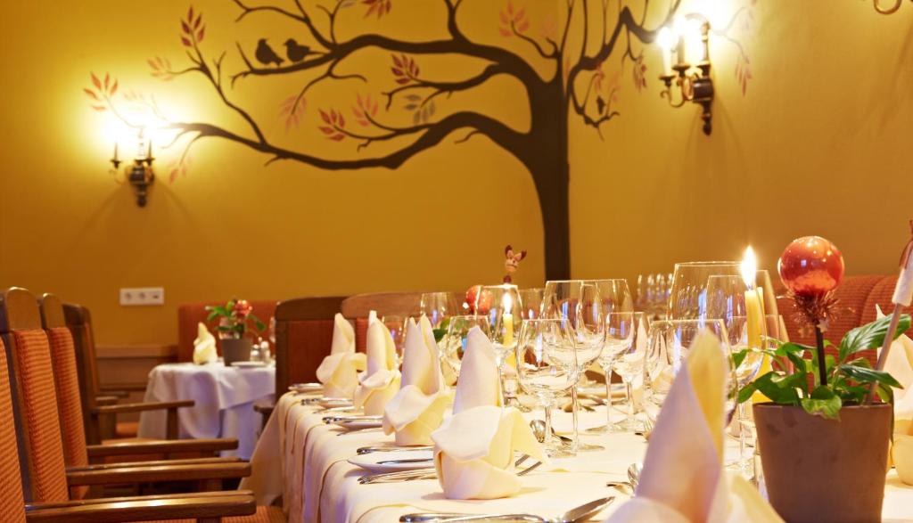 Eleganti iekārtota restorāna galda skats ar glāzēm, salvetēm un ziediem, fonā redzama sienas glezna ar koku un putniem. Atmosfēra ir silta un viesmīlīga.