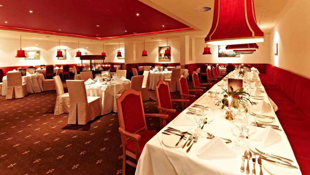 Eleganta restorāna iekārtojums ar sarkaniem akcentiem un svinīgu galdu klājumu, kas izveidots romantiskai vai svinīgai atmosfērai. Sarkanās lampas un izsmalcināti galda piederumi papildina kopējo noskaņu.