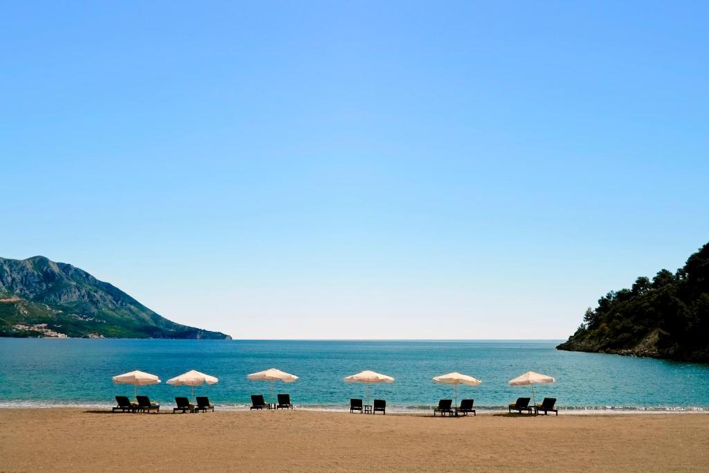 Pludmales ainava ar sauļošanās krēsliem un lietussargiem, kas izvietoti gar krastu, ar kalnu fona un skaidru debesu fonu.