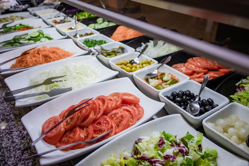 Dažādu salātu sastāvdaļu bārs ar tomātiem, gurķiem, sīpoliem un citiem dārzeņiem, kas sakārtoti baltās traukos.