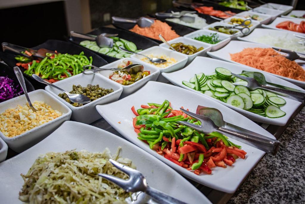 Dažādas svaigas dārzeņu salātu sastāvdaļas, tostarp gurķi, paprika, burkāni un sīpoli, pasniegtas uz baltiem šķīvjiem.