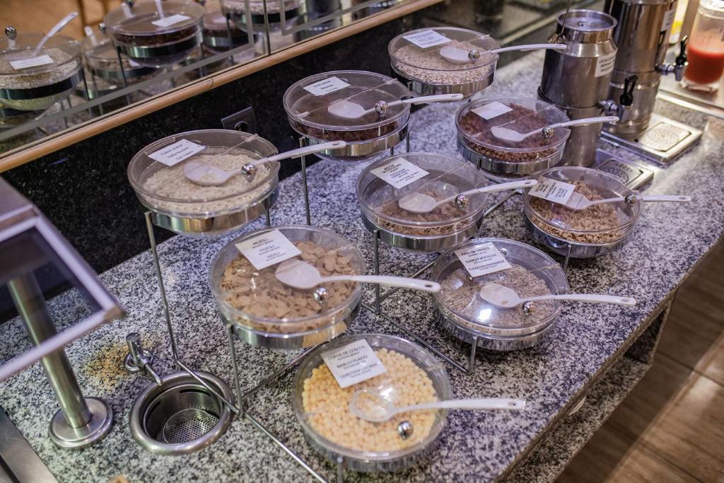 Dažādas graudaugu un pārslu izvēles izkārtotas stikla traukos uz galda, ar etiķetēm un karotēm. Ideāli piemērots brokastu bufetei.