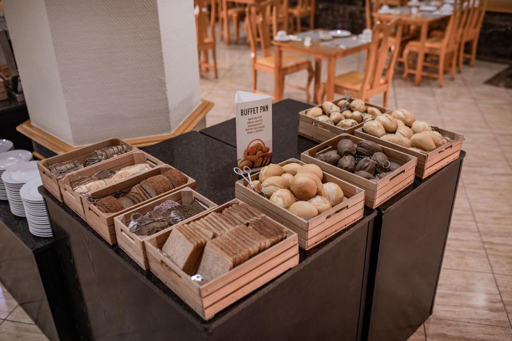 Piedāvājums ar dažādiem maizes veidiem un maizītēm, sakārtots koka kastēs. Uz galda redzams arī informatīvs stends par bufeti.