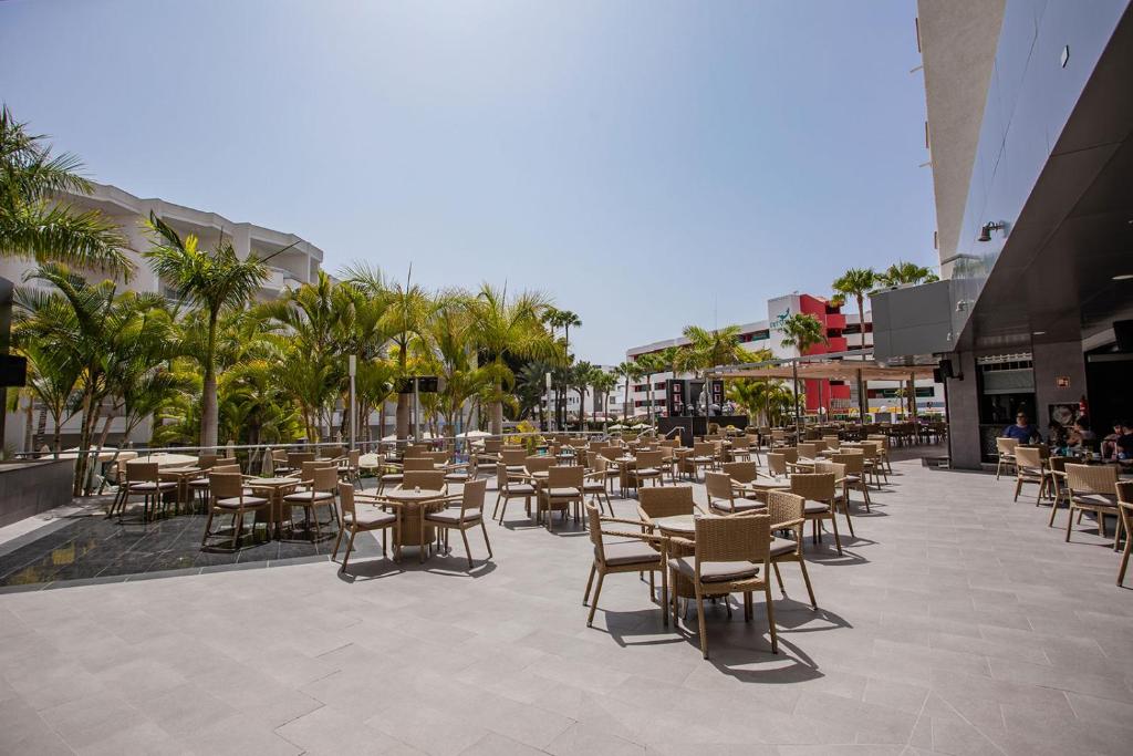 Ārtelpa ar daudziem galdiem un krēsliem, ko ieskauj palmas, ideāla vieta relaksācijai un ēšanai saulainā dienā. Moderns restorāns ar plašu terasi.