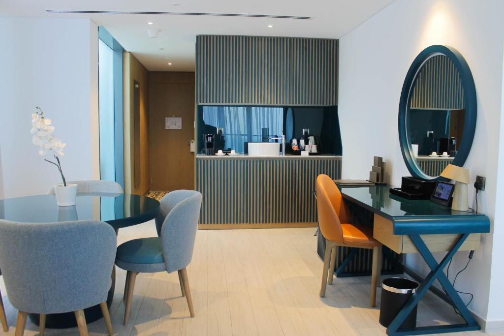 Mūsdienīgs viesnīcas numurs ar elegantu interjeru, ietverot galdu ar krēsliem, darba staciju un mini bāru. Interjers ir gaišs un stilīgs, radot komfortablu atmosfēru.