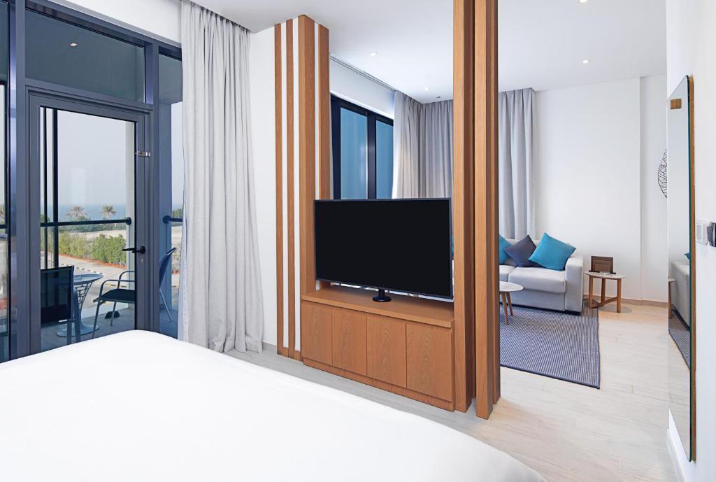 Mūsdienīgs viesnīcas numurs ar atvērtu plānojumu, kurā redzams televizors un komfortabla sēdvieta, kā arī skats uz terasi un ainavu.